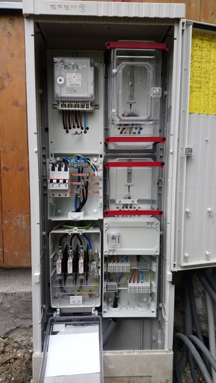 Otwarta skrzynka elektryczna z licznikiem, bezpiecznikami i różnymi złączami kablowymi, widoczne kable wchodzące do skrzynki od dołu.
