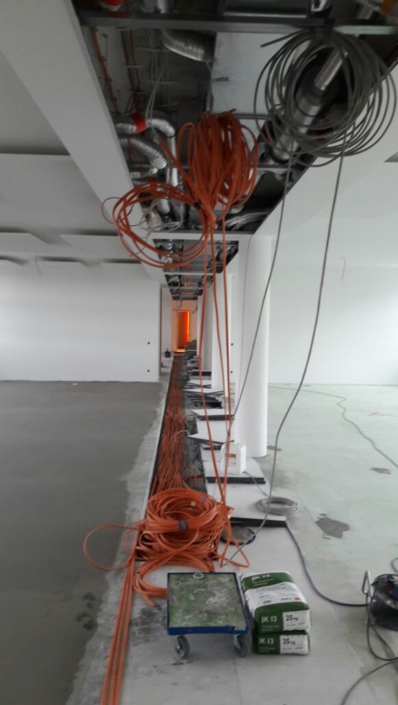 Pomarańczowe i szare kable zwisające z sufitu podwieszanego w trakcie instalacji w nowym budynku, widoczne worki z zaprawą budowlaną i wózek transportowy.