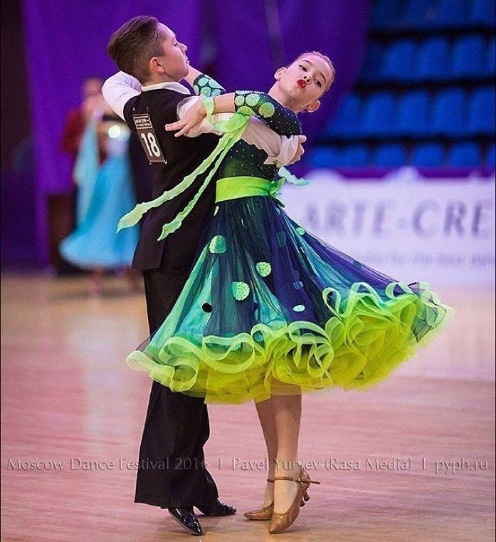 Młoda para taneczna w strojach galowych podczas tańca towarzyskiego. Dziewczyna w sukience z falbanami w odcieniach zieleni i granatu, chłopak w czarnym garniturze z numerem startowym 18.