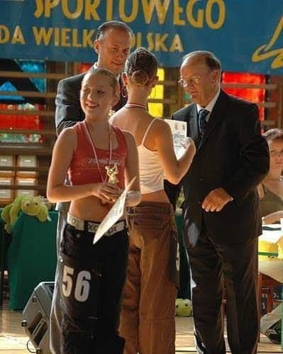 Młoda tancerka z numerem 56 odbiera nagrodę na turnieju tańca towarzyskiego, wręczana przez sędziów w garniturach, na tle baneru sportowego.