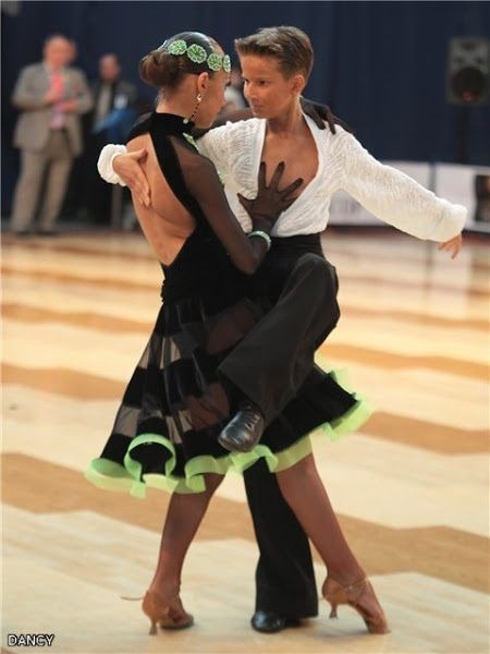 Para młodych tancerzy w strojach turniejowych podczas tańca towarzyskiego: dziewczyna w czarnej sukience z zielonymi akcentami, chłopak w białej koszuli i czarnych spodniach, na parkiecie tanecznym.