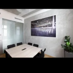 Sala konferencyjna z teksturowanymi szarymi ścianami, dużym obrazem hali przemysłowej, stołem z czarnymi krzesłami i rośliną w donicy.