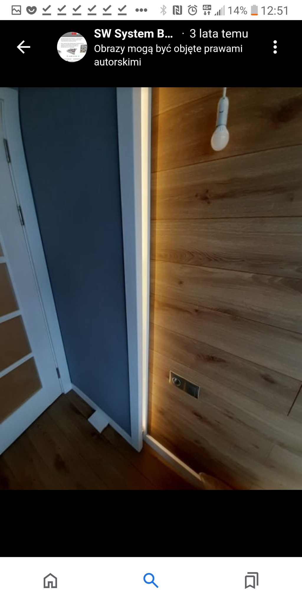 Wykończone wnętrze z drewnianą okładziną ścienną, podświetleniem LED wzdłuż krawędzi ściany i szarą ścianą, widoczne gniazdko elektryczne.