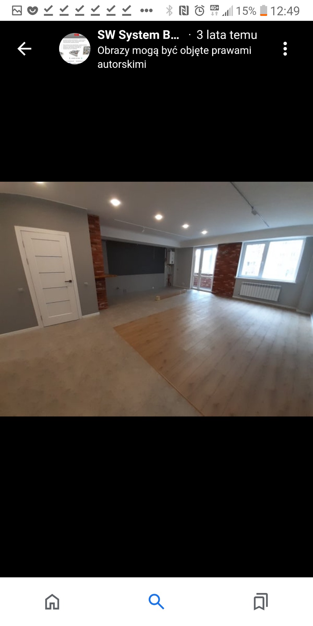 Wnętrze pokoju w trakcie prac wykończeniowych: położona częściowo podłoga z paneli, pomalowane szare ściany, białe drzwi, fragment ściany z cegły, biały grzejnik pod oknem.