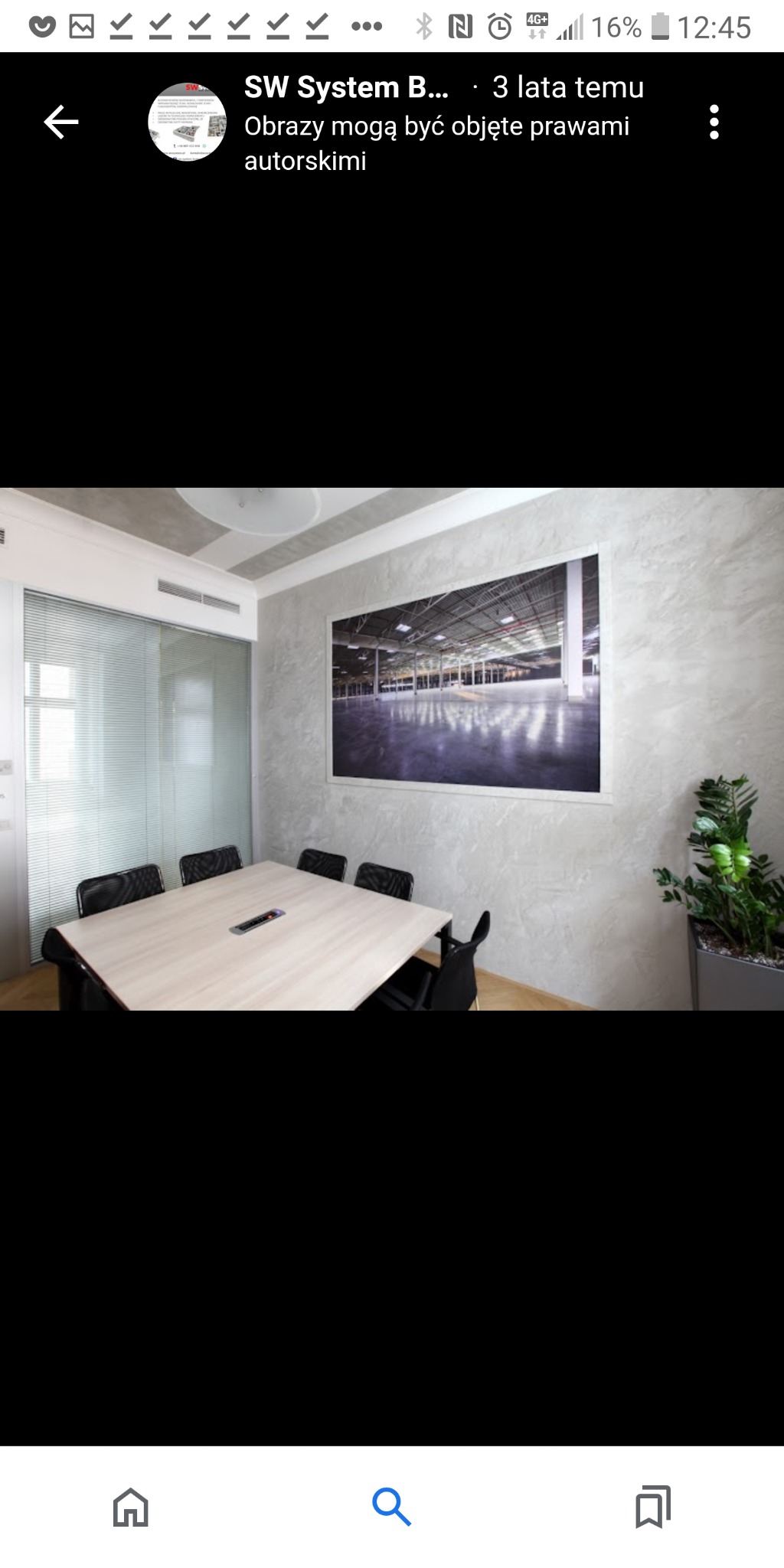 Sala konferencyjna z teksturowanymi szarymi ścianami, dużym obrazem hali przemysłowej, stołem z czarnymi krzesłami i rośliną w donicy.