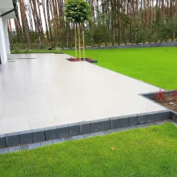 Nowoczesny taras z jasnych płyt, obrzeża z ciemnej kostki, trawnik i drzewa w tle, minimalistyczny design.