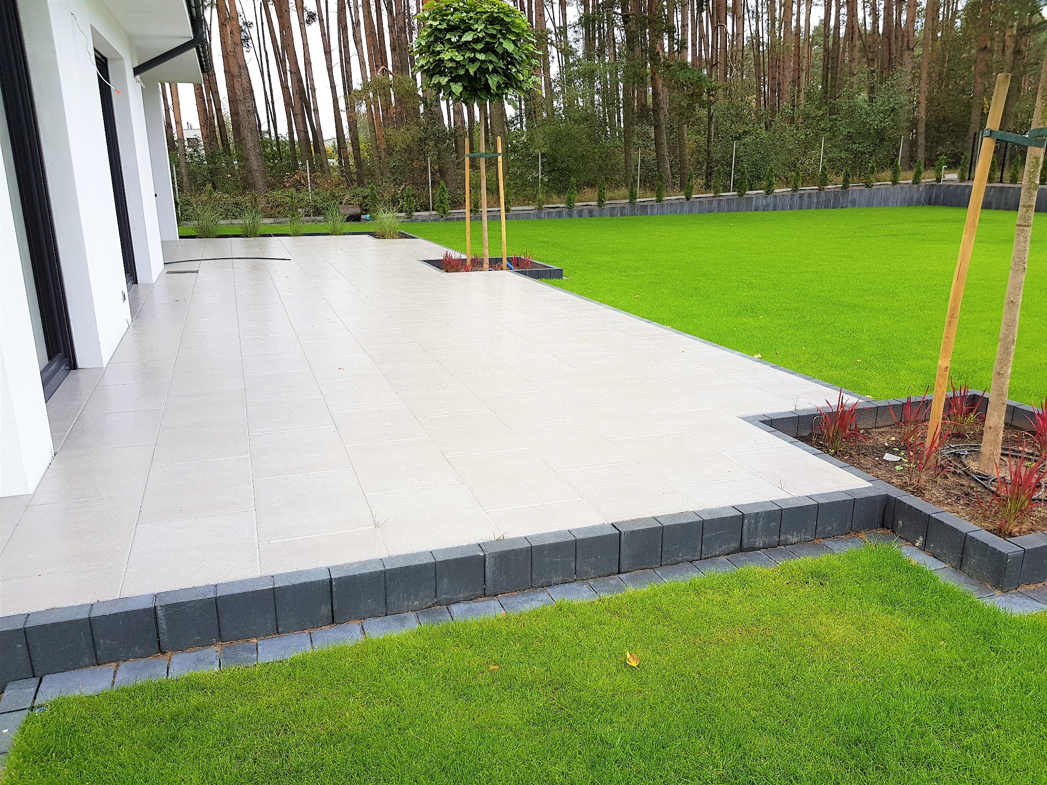 Nowoczesny taras z jasnych płyt, obrzeża z ciemnej kostki, trawnik i drzewa w tle, minimalistyczny design.