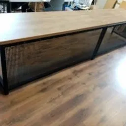 Stół z blatem imitującym drewno i metalową, ażurową konstrukcją, widoczne spawane elementy ramy, na podłodze z paneli.