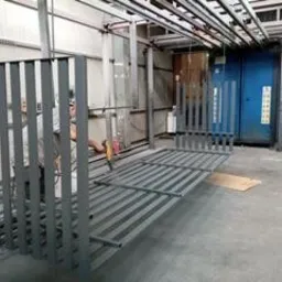 Szare metalowe elementy konstrukcyjne, prawdopodobnie fragmenty ogrodzenia lub balustrady, ułożone na podłodze hali produkcyjnej. W tle widoczny pracownik z narzędziem oraz niebieska komora...