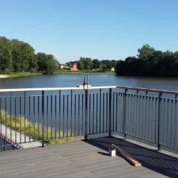 Nowoczesna balustrada z metalowymi pionowymi prętami i drewnianym poręczą, zamontowana na tarasie z widokiem na jezioro w słoneczny dzień, narzędzia widoczne na podłodze.