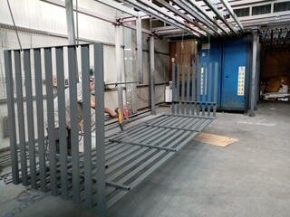 Szare metalowe elementy konstrukcyjne, prawdopodobnie fragmenty ogrodzenia lub balustrady, ułożone na podłodze hali produkcyjnej. W tle widoczny pracownik z narzędziem oraz niebieska komora...