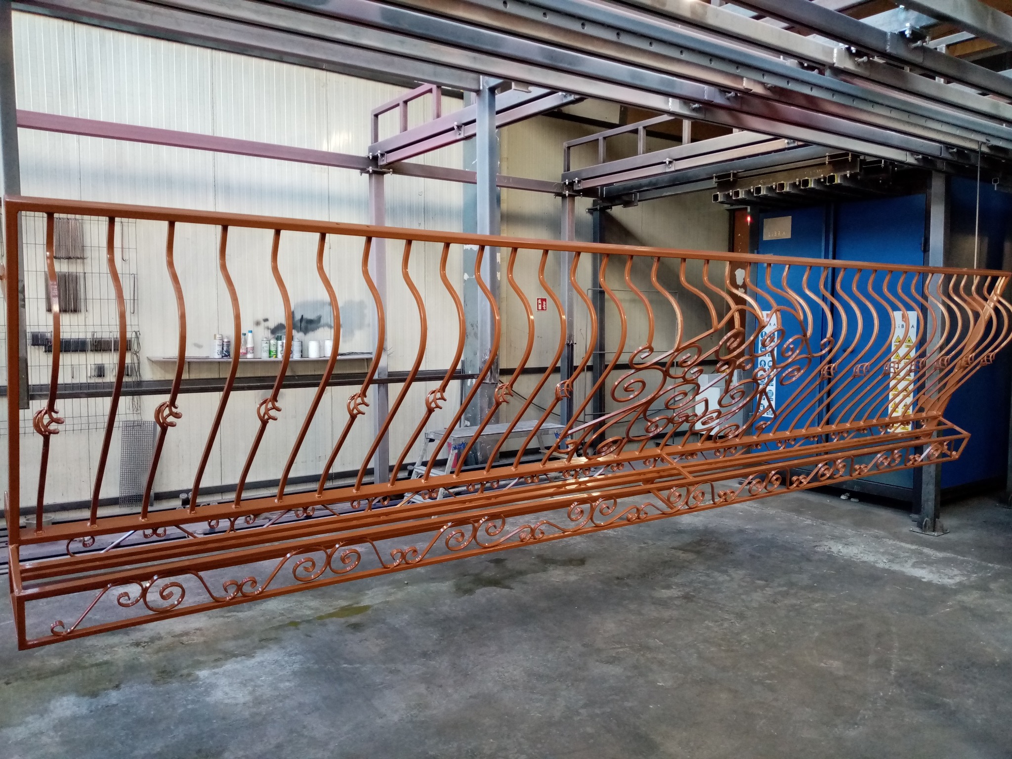 Element balustrady z metalu w kolorze miedzianym, z dekoracyjnymi, spiralnymi zdobieniami, zawieszony na konstrukcji w hali produkcyjnej, w tle widoczne narzędzia i niebieska szafa.