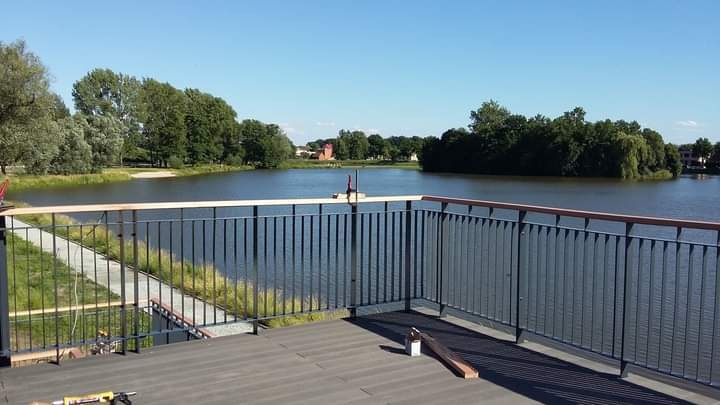 Nowoczesna balustrada z metalowymi pionowymi prętami i drewnianym poręczą, zamontowana na tarasie z widokiem na jezioro w słoneczny dzień, narzędzia widoczne na podłodze.