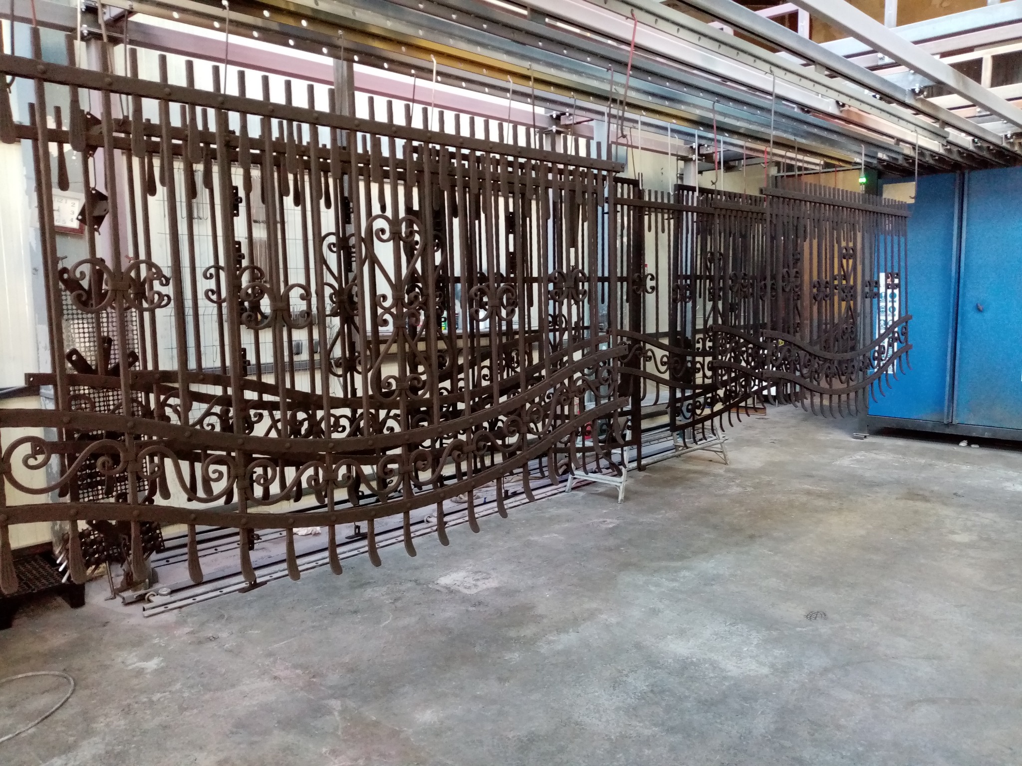Metalowe elementy ogrodzenia z dekoracyjnymi wzorami, zawieszone na szynach w hali produkcyjnej, przygotowane do malowania lub obróbki.
