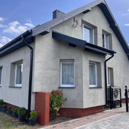 KONRAD FIEREK FROSEN - Elewacja domu jednorodzinnego z oknami PCV, wykończona tynkiem o fakturze baranka. Widoczne czarne rynny, ceglana podmurówka i brama w tle. Na ścianie kosz do koszykówki.