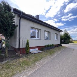 KONRAD FIEREK FROSEN - Elewacja domu jednorodzinnego z oknami PCV, widok z boku na szarą fasadę, czarne rynny i fragment ogrodzenia. Błękitne niebo z chmurami w tle.