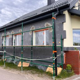 KONRAD FIEREK FROSEN - Dom w trakcie ocieplania: szare płyty termoizolacyjne na elewacji, rusztowanie z zielonych rur, okna zabezpieczone folią. Fragment elewacji w kolorze kremowym.