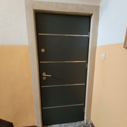 KONRAD FIEREK FROSEN - Ciemne drzwi wejściowe z poziomymi, jasnymi paskami i ościeżnicą w odcieniach beżu, zamontowane w bloku mieszkalnym z pomarańczowymi ścianami.