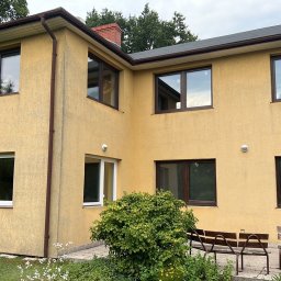 KONRAD FIEREK FROSEN - Żółta elewacja domu z brązowymi oknami PCV i rynnami, widoczny fragment ogrodu z zielenią i meblami ogrodowymi. Dom z dwoma kondygnacjami.