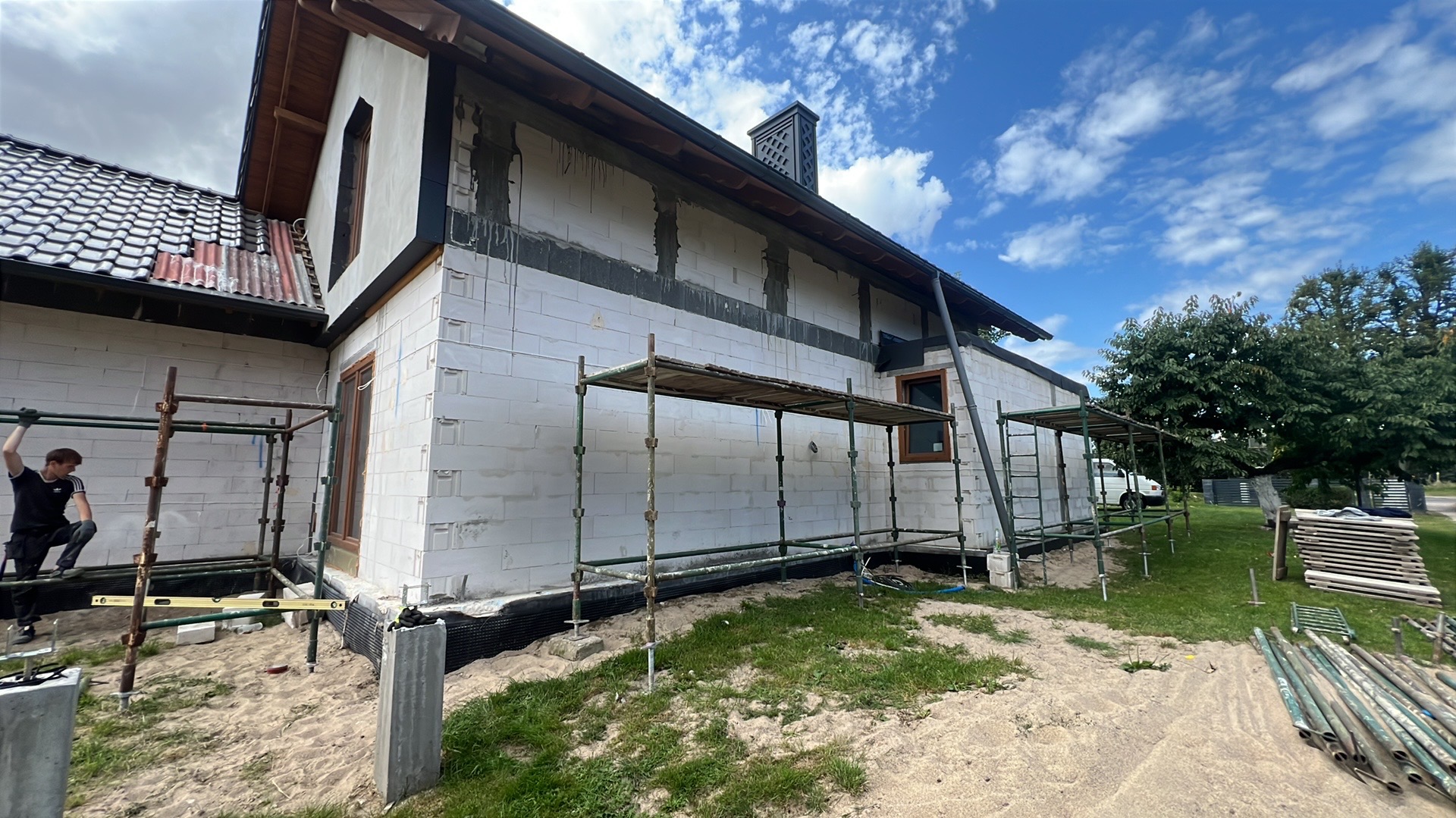 Dom w trakcie ocieplania: białe bloczki, rusztowania, okna. Widoczny fragment dachu z uszkodzonymi dachówkami. Na pierwszym planie piasek i trawa.