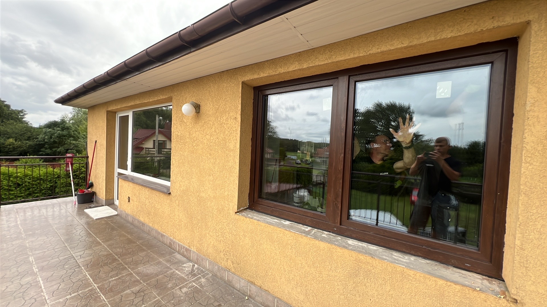 Żółta elewacja domu z nowymi oknami PCV z brązowymi ramami, widok z zewnątrz balkonu. W oknach odbijają się osoby robiące zdjęcie.