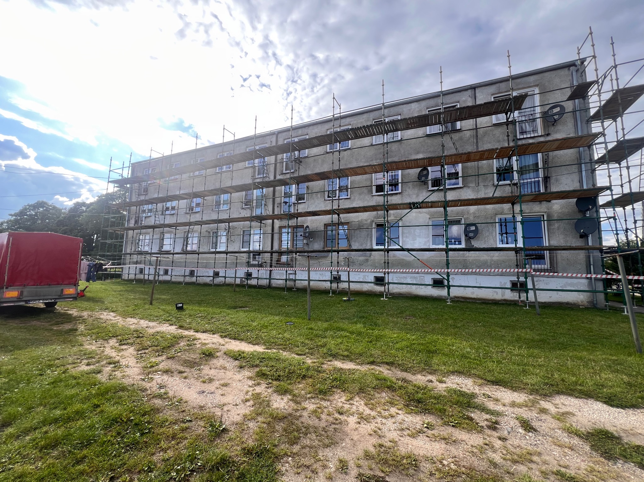 Budynek wielorodzinny w trakcie prac elewacyjnych, otoczony rusztowaniami. Widoczny fragment przyczepy z plandeką. Ujęcie w słoneczny dzień z perspektywy trawnika.