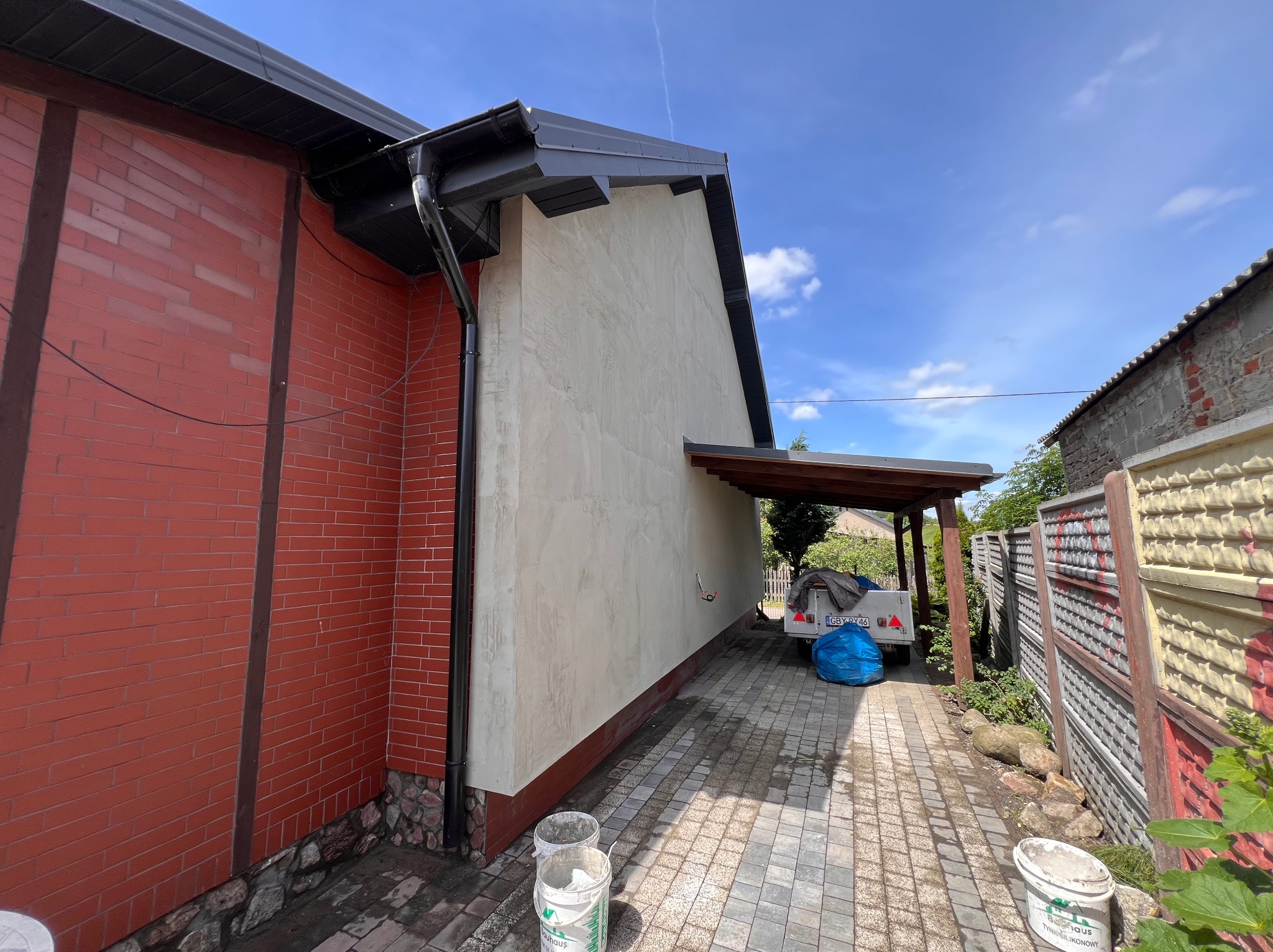 Elewacja domu po tynkowaniu, z widocznymi detalami tekstury. Część ściany wykończona czerwoną cegłą, obok zaparkowana przyczepka. Widoczne wiaderka po tynku.