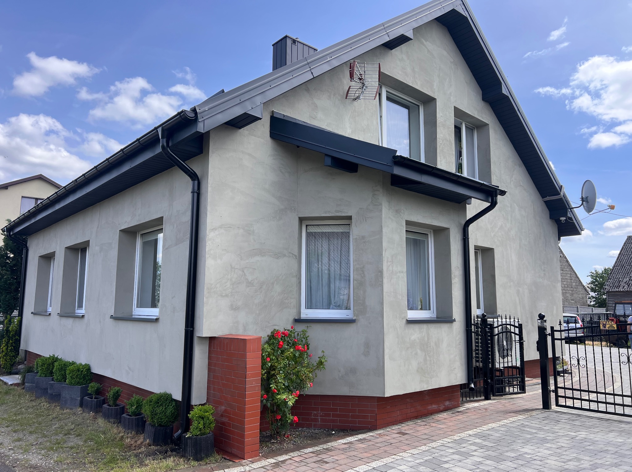 Elewacja domu jednorodzinnego z oknami PCV, wykończona tynkiem o fakturze baranka. Widoczne czarne rynny, ceglana podmurówka i brama w tle. Na ścianie kosz do koszykówki.