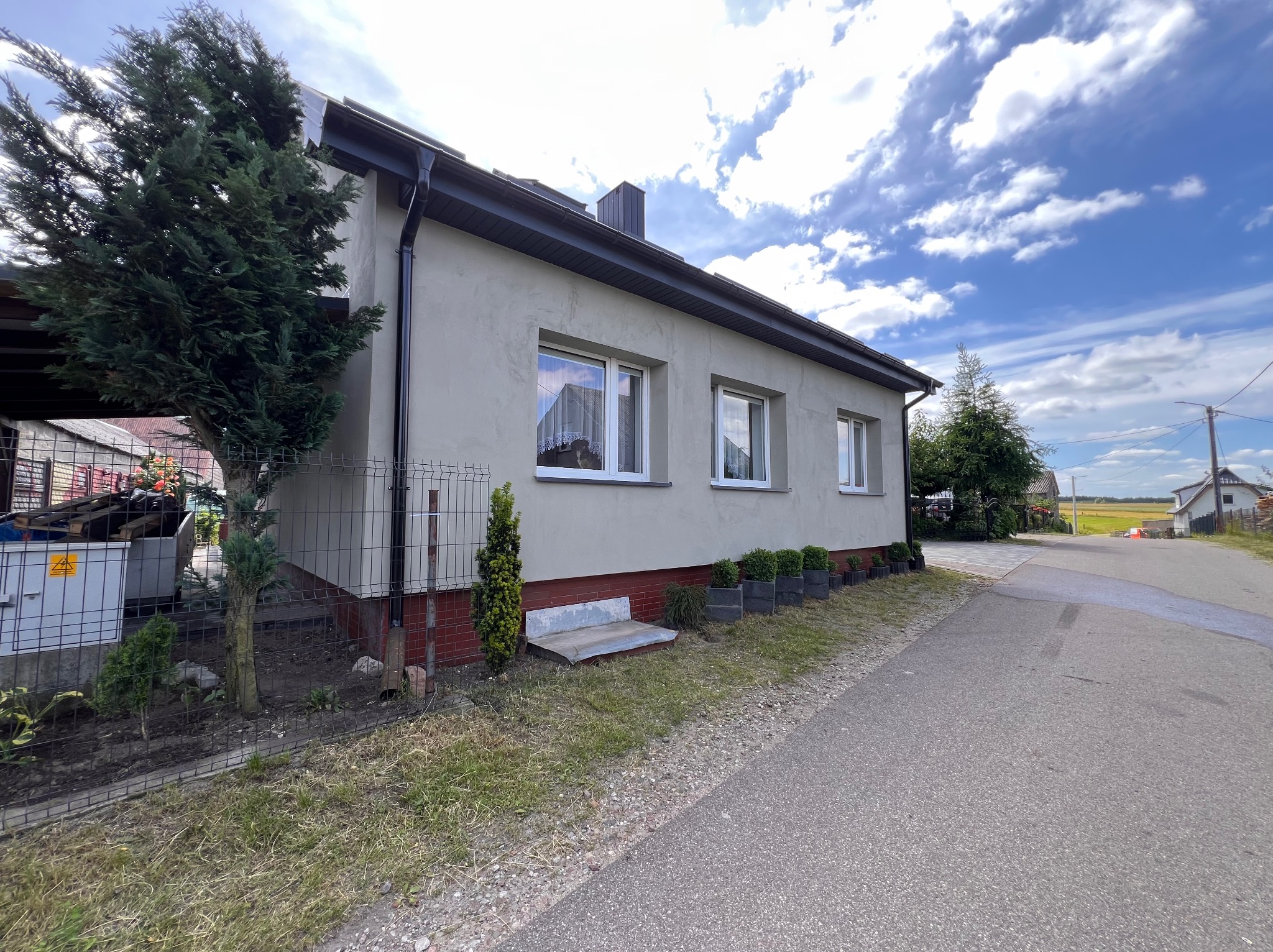 Elewacja domu jednorodzinnego z oknami PCV, widok z boku na szarą fasadę, czarne rynny i fragment ogrodzenia. Błękitne niebo z chmurami w tle.