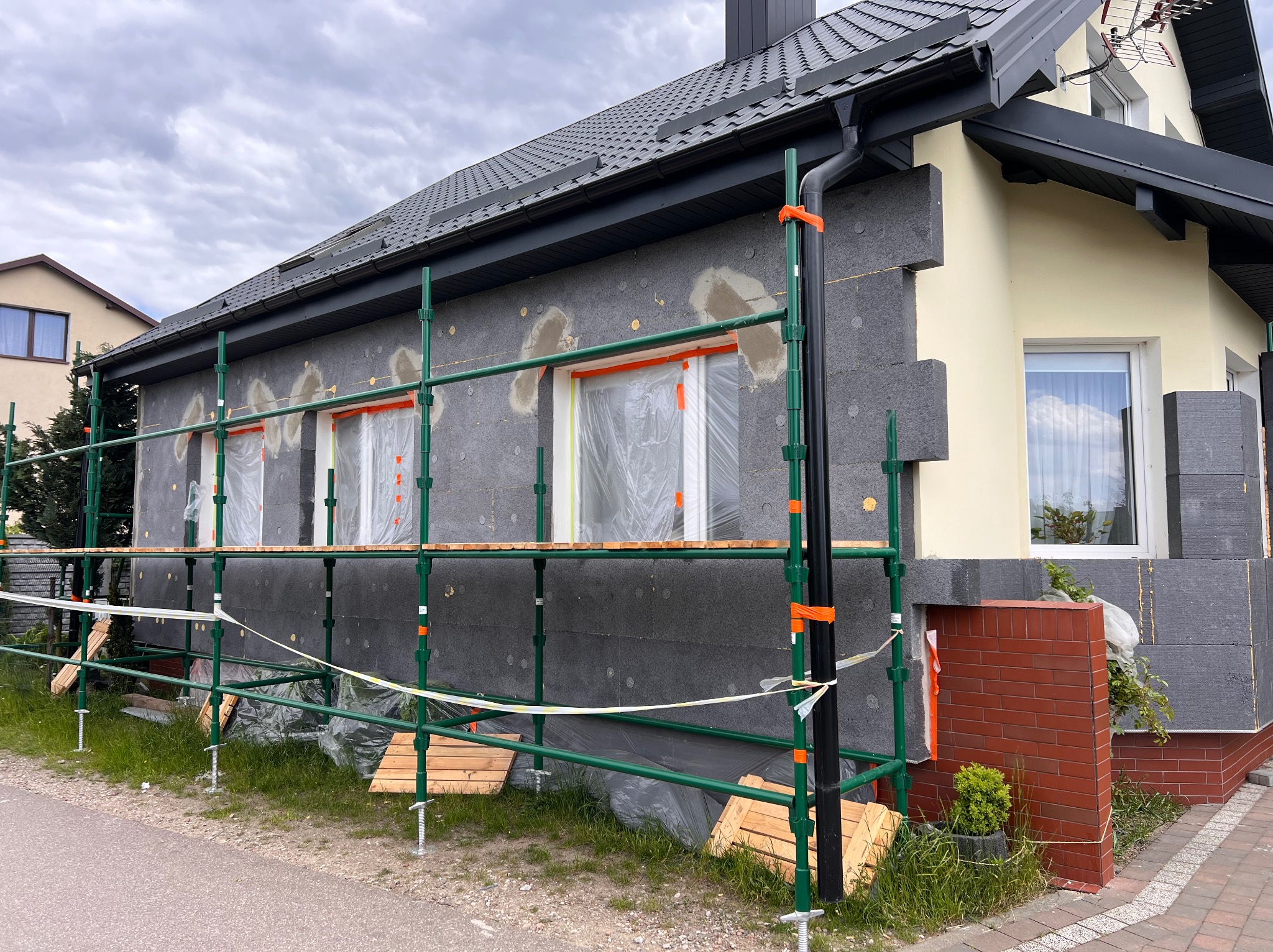 Dom w trakcie ocieplania: szare płyty termoizolacyjne na elewacji, rusztowanie z zielonych rur, okna zabezpieczone folią. Fragment elewacji w kolorze kremowym.