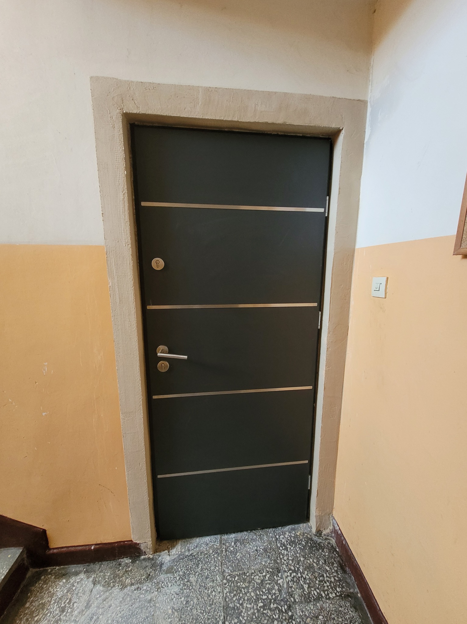 Ciemne drzwi wejściowe z poziomymi, jasnymi paskami i ościeżnicą w odcieniach beżu, zamontowane w bloku mieszkalnym z pomarańczowymi ścianami.