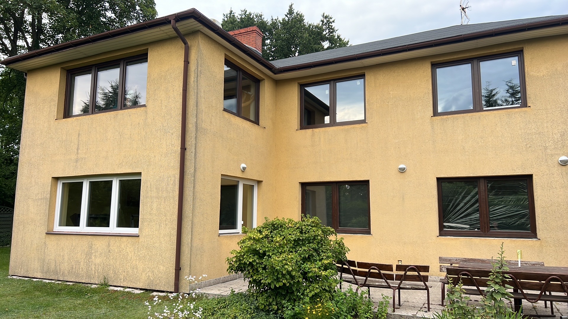 Żółta elewacja domu z brązowymi oknami PCV i rynnami, widoczny fragment ogrodu z zielenią i meblami ogrodowymi. Dom z dwoma kondygnacjami.