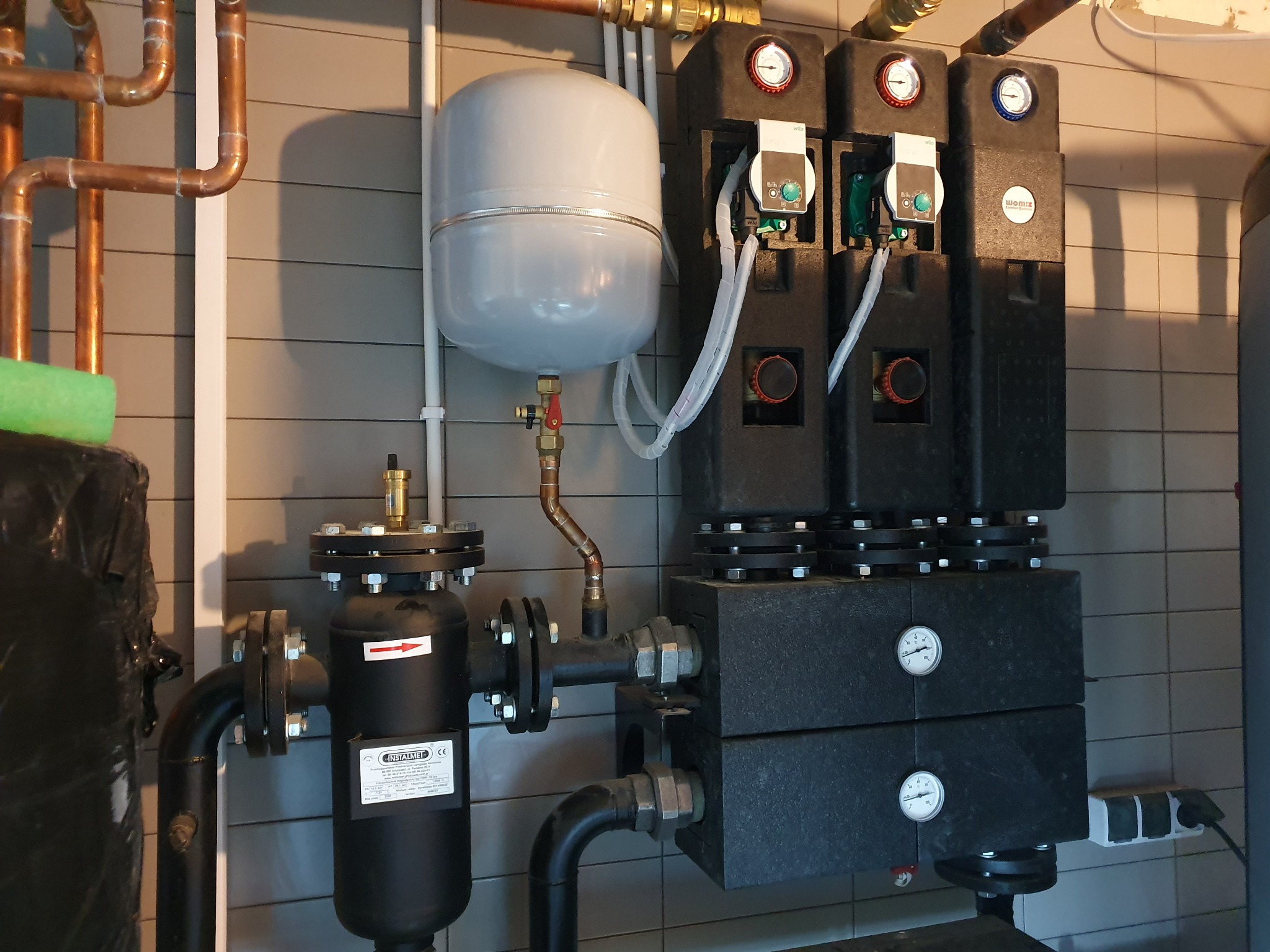Zamknięty system grzewczy z pompami obiegowymi, zbiornikiem wyrównawczym i filtrem, zainstalowany przy szarej, kafelkowanej ścianie.