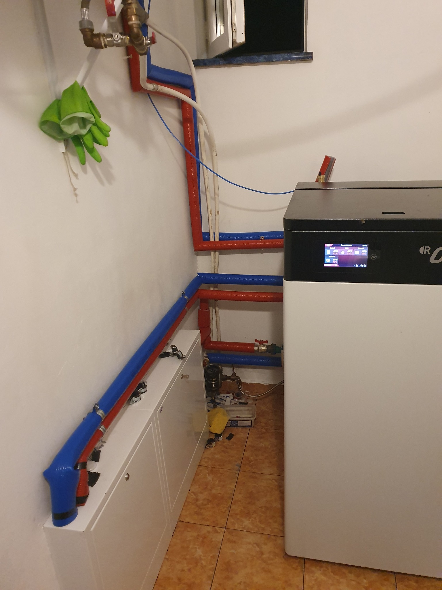 Nowoczesny piec grzewczy z wyświetlaczem, podłączony do instalacji z rur w kolorach czerwonym i niebieskim, zielone rękawice robocze wiszą na ścianie, widoczne białe szafki instalacyjne.