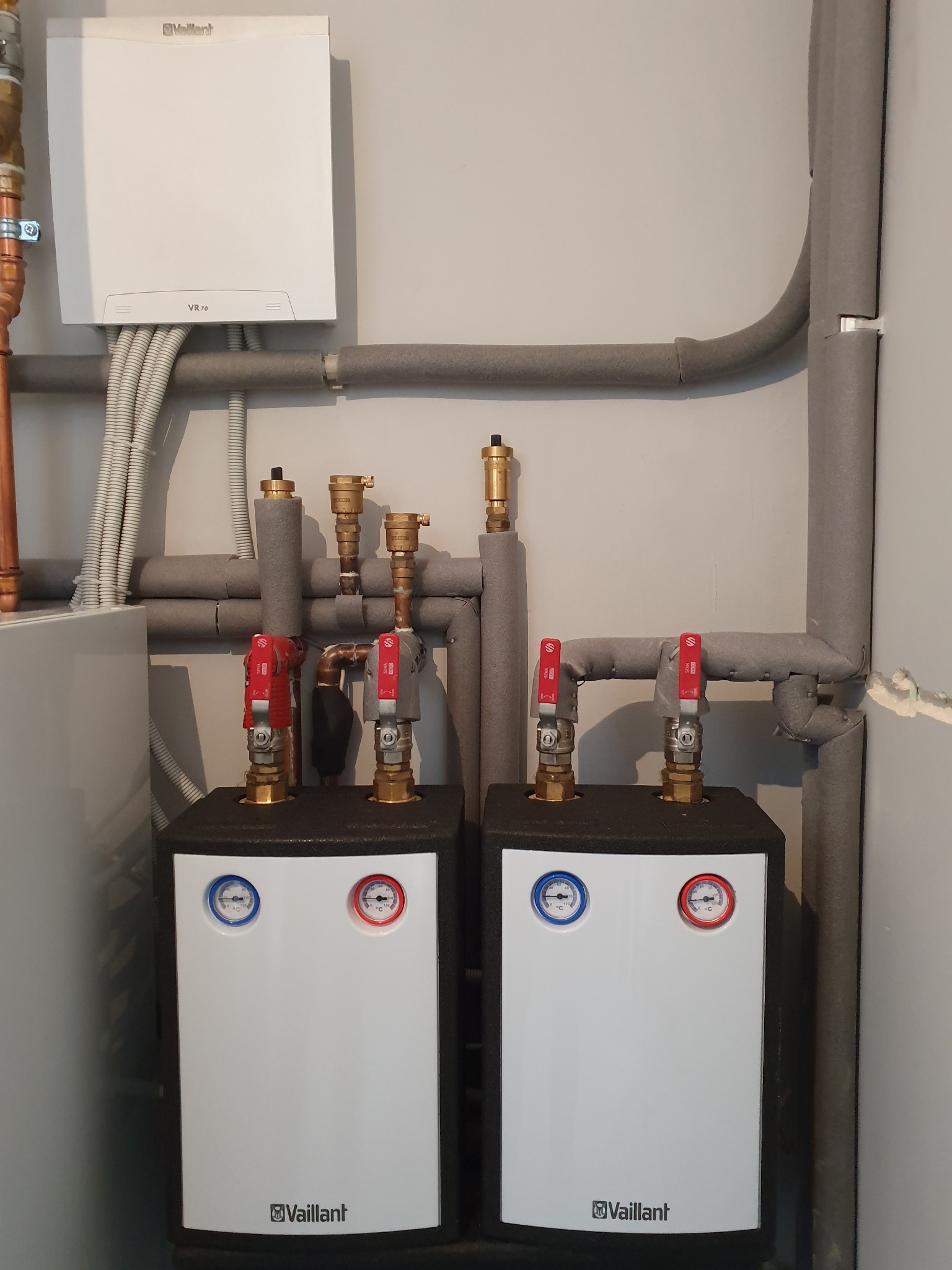 Dwa białe kotły grzewcze Vaillant z manometrami, połączone rurami w izolacji termicznej, zamontowane na szarej ścianie. Widoczny również moduł sterujący Vaillant VR 70.
