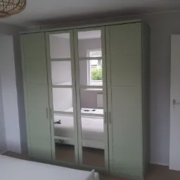 Jasnozielona szafa z lustrami wstawionymi w drzwi, odbijająca wnętrze pokoju z łóżkiem i oknem, ustawiona na tle szarej ściany i białych drzwi.