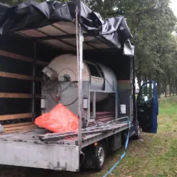 Ciężki, cylindryczny zbiornik przemysłowy zabezpieczony plandeką w przestrzeni ładunkowej otwartego busa na tle drzew.