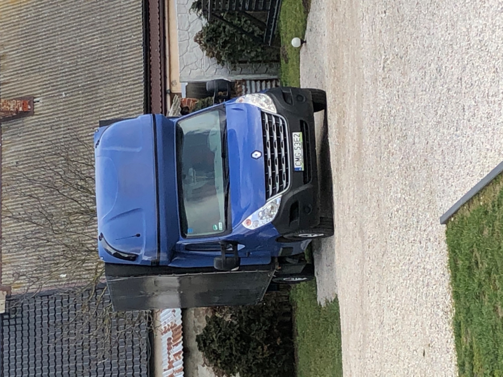 Niebieski samochód dostawczy Renault Master z czarną skrzynią ładunkową, zaparkowany na żwirowej drodze przed budynkami z różnymi rodzajami dachów.