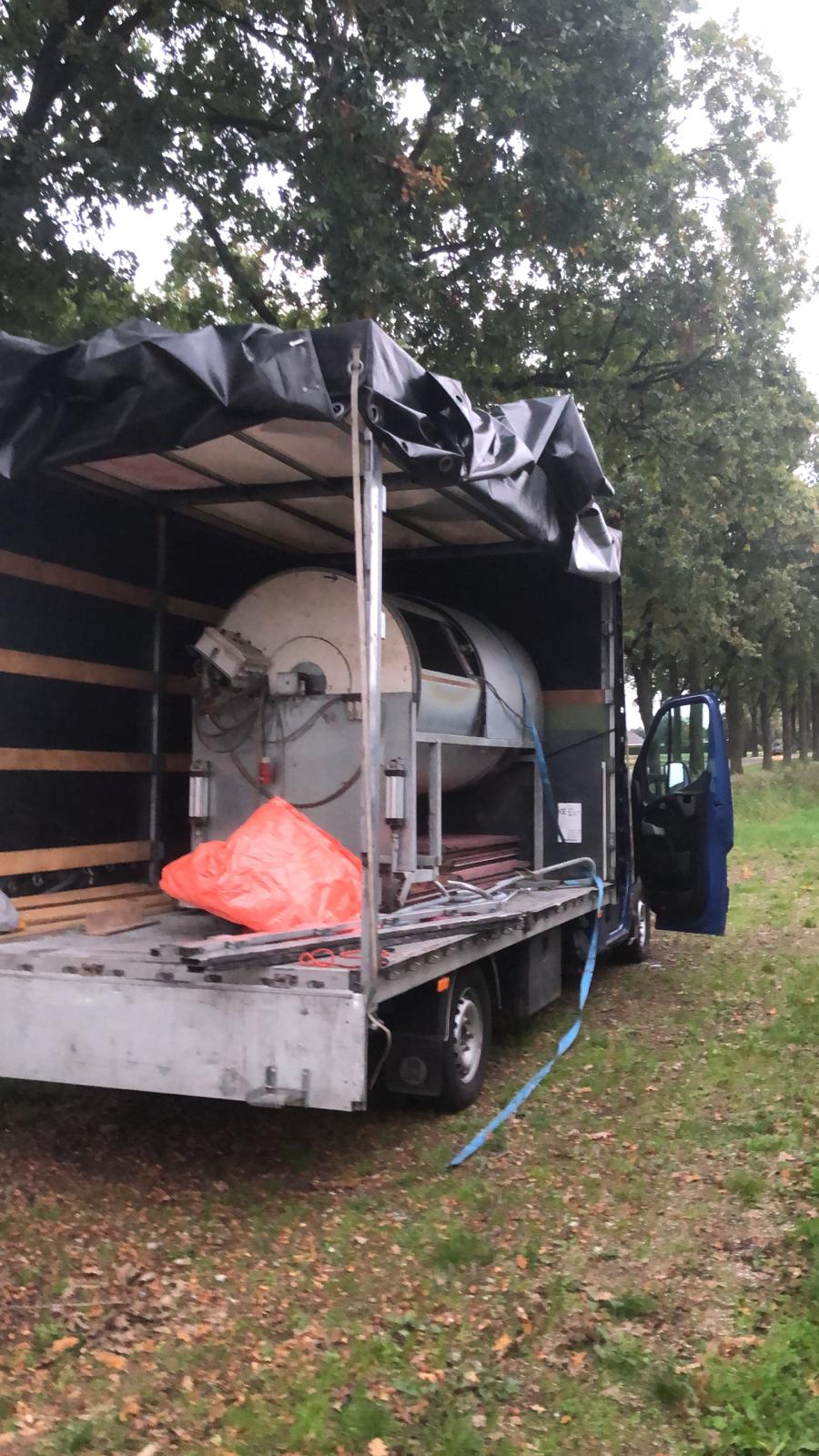 Ciężki, cylindryczny zbiornik przemysłowy zabezpieczony plandeką w przestrzeni ładunkowej otwartego busa na tle drzew.