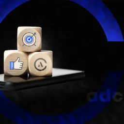 Trzy drewniane kostki z ikonami celu, polubienia i litery A, ułożone na smartfonie na tle ciemnego, teksturowanego tła z niebieskim okręgiem i napisem adcube.