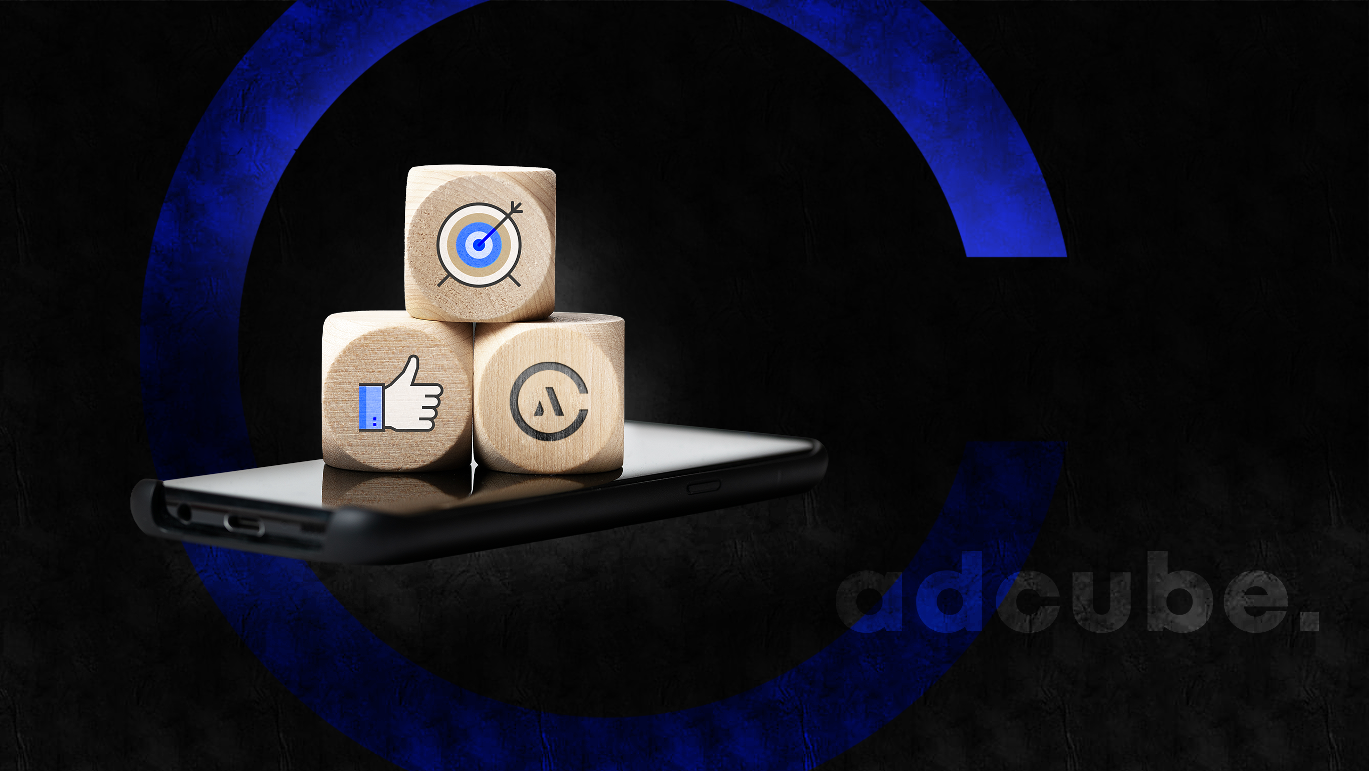 Trzy drewniane kostki z ikonami celu, polubienia i litery A, ułożone na smartfonie na tle ciemnego, teksturowanego tła z niebieskim okręgiem i napisem adcube.