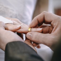 Foto Studio Andrzej Barełkowski - Zakładanie obrączki podczas ceremonii ślubnej we Wschowie. Dłonie w zbliżeniu, widoczna złota obrączka i fragment sukni panny młodej. Delikatne światło.