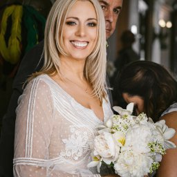 Foto Studio Andrzej Barełkowski - Uśmiechnięta panna młoda w koronkowej sukni, trzymająca bukiet białych kwiatów, z mężczyzną w tle. Delikatne światło podkreśla detale sukni i makijaż.