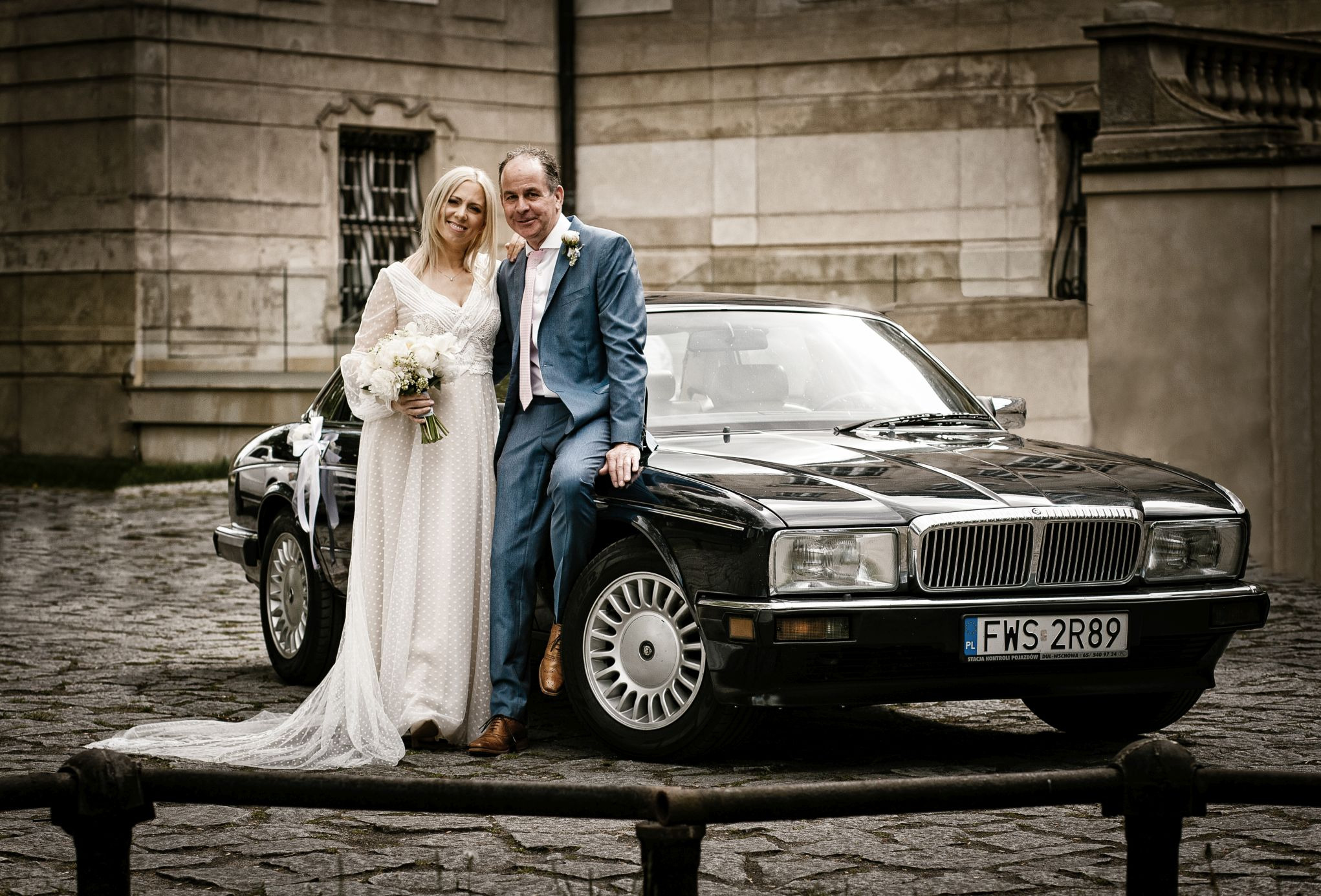 Para młoda w strojach ślubnych pozuje przy czarnym samochodzie Jaguar na tle kamiennej architektury, panna młoda trzyma bukiet białych kwiatów.