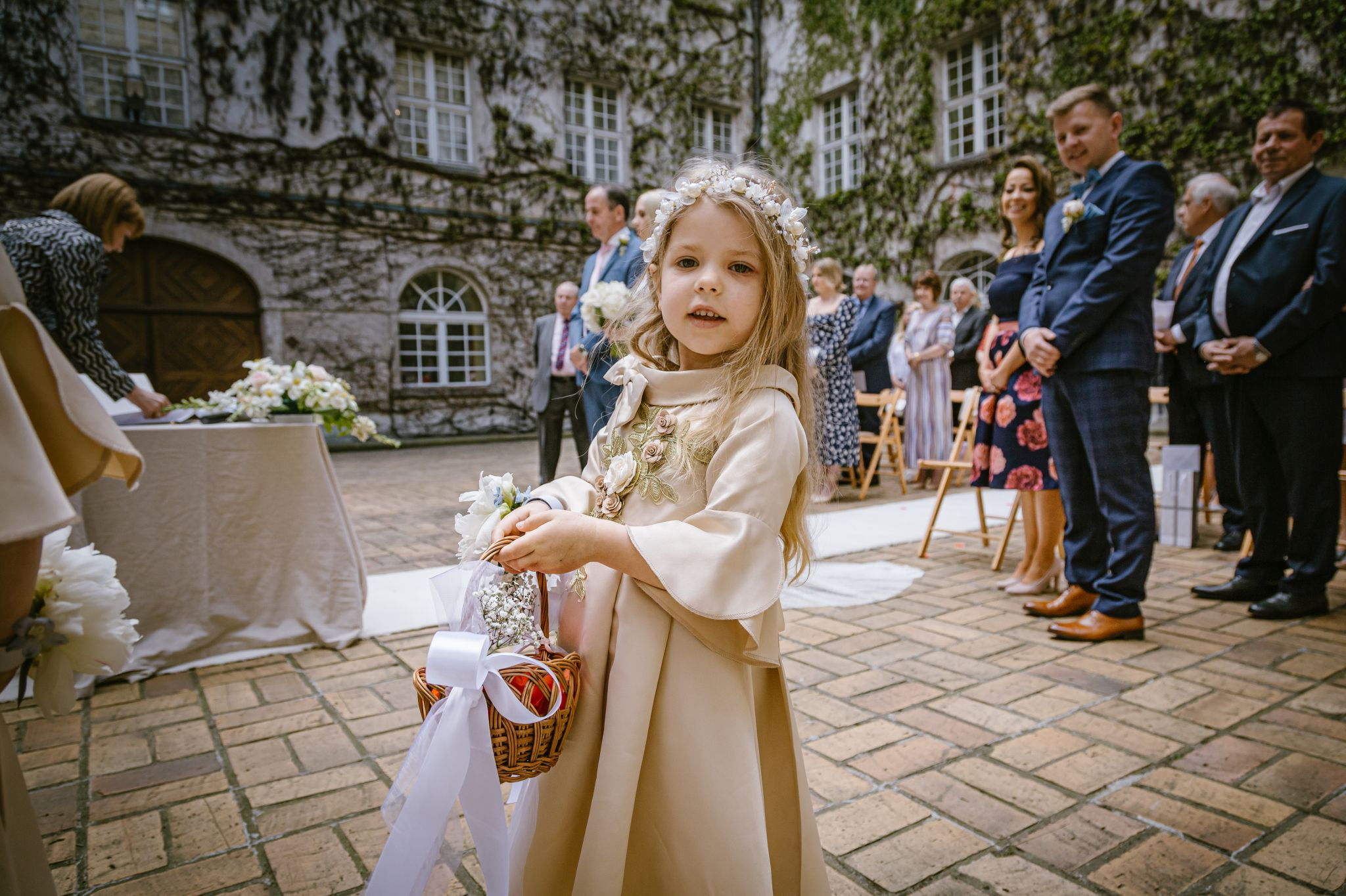 Dziewczynka w beżowej sukience i wianku na głowie, niosąca koszyczek z kwiatami podczas ceremonii ślubnej na tle kamiennej ściany porośniętej bluszczem.
