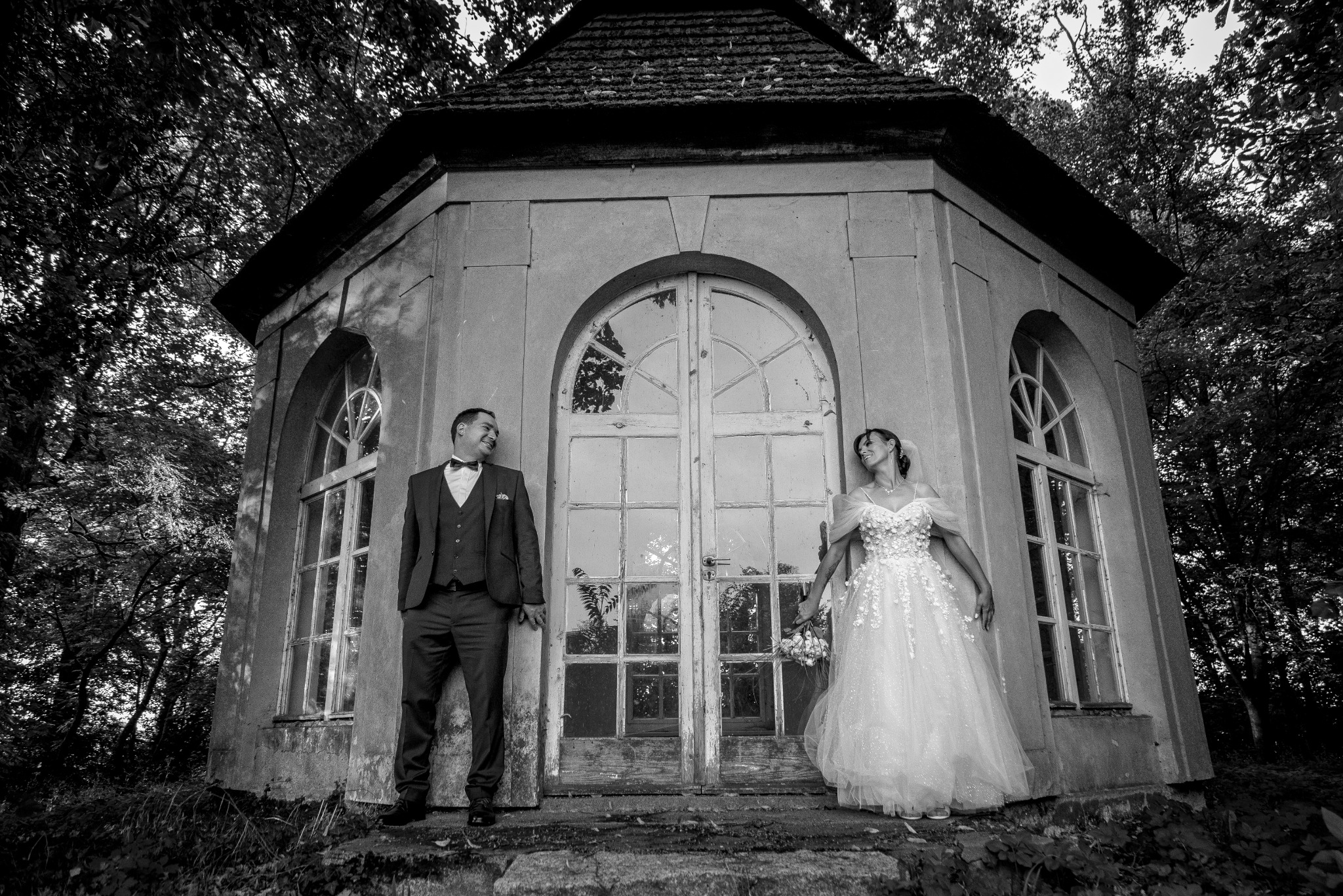 Czarno-białe zdjęcie pary młodej pozującej przy zabytkowej, ośmiokątnej altanie z dużymi, łukowatymi oknami; pan młody w ciemnym garniturze z muszką, panna młoda w białej sukni z aplikacjami...