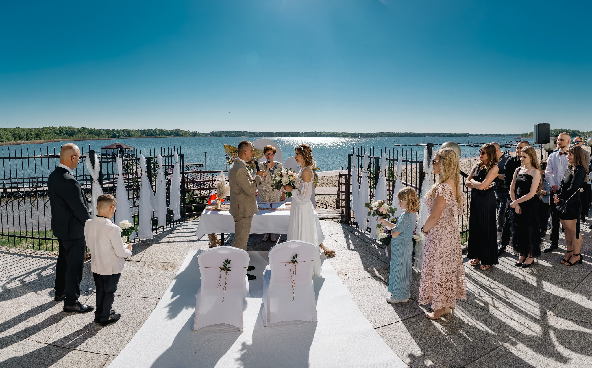 Plenerowa ceremonia ślubna nad jeziorem: para młoda, goście, dekoracje w bieli, słoneczny dzień. Ujęcie z szerokim kadrem, podkreślające scenerię.