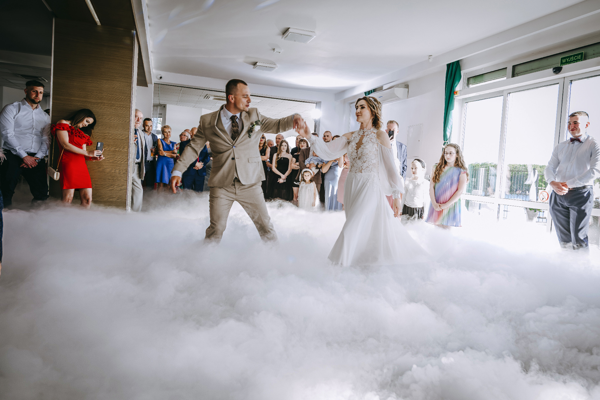Para młoda w tańcu na 'chmurach' podczas wesela. Pan w beżowym garniturze, pani w białej sukni. Goście weselni w tle. Efekt dymu na parkiecie.