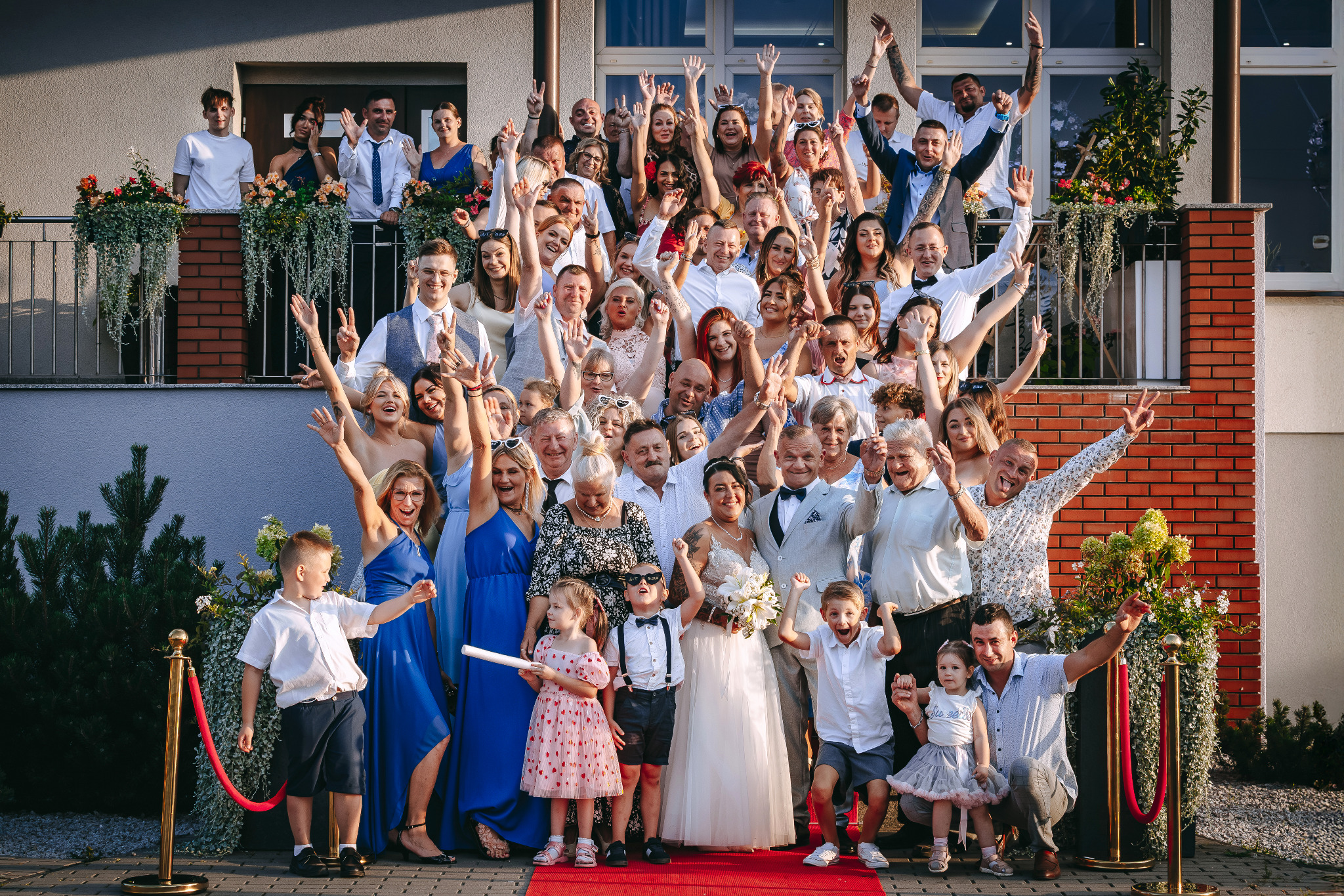 Grupowe zdjęcie weselne z panną młodą i panem młodym na czerwonym dywanie, goście z podniesionymi rękami, balkon z kwiatami w tle, radosna atmosfera.