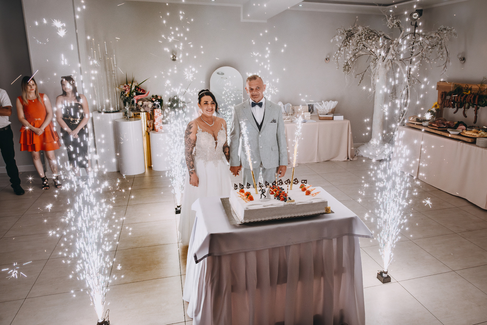 Uśmiechnięta para młoda trzymająca się za ręce za tortem weselnym, otoczona fontannami iskier w jasnym wnętrzu sali bankietowej. Dekoracje i goście w tle.
